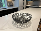 May include: A black, decorative fruit bowl with a unique openwork design. The bowl is round with a wide, shallow shape and sits on a white countertop. The bowl's intricate pattern creates a shadow on the surface below. The kitchen features a window, cabinets, and a stainless steel faucet.