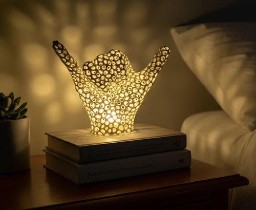 May include: A decorative lamp shaped like a hand with the 'hang loose' gesture. The lamp is made of a porous material, allowing light to shine through. It sits on top of two books on a wooden nightstand. The lamp emits a warm, inviting glow.