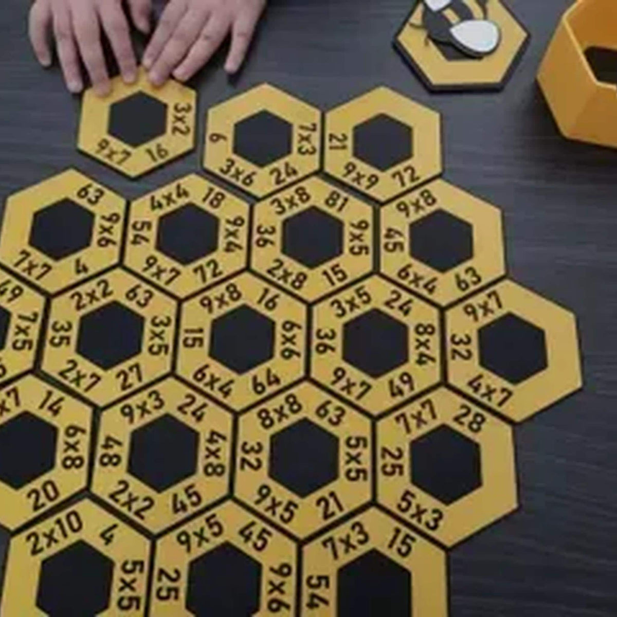 Child's hands positioning a yellow multiplication tile among the separated HoneyHex pieces on a dark wood table.