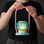Hands holding a Coral Cove lantern with pink handle, teal ribbed shade, yellow center and pale blue base against a dark shirt.