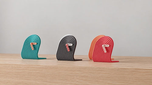 Angled close-up of Archwave Sculptural Desk Clock examples in contrasting colors on light oak against a gray wall.
