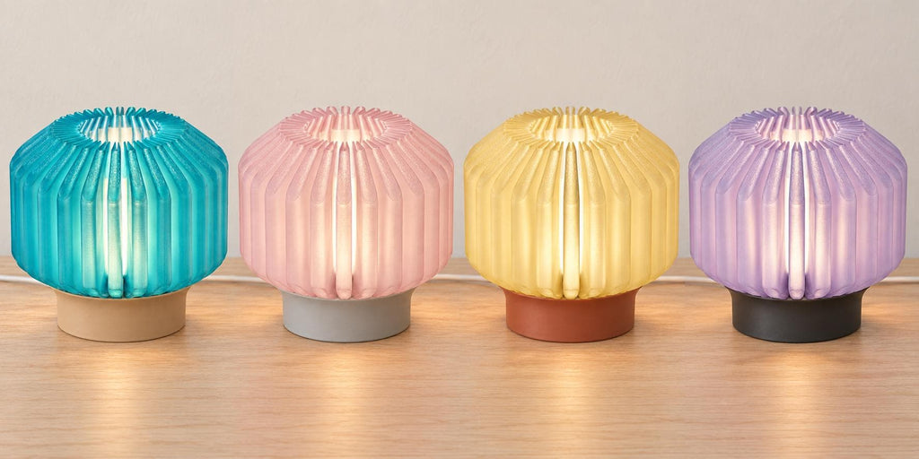 Four illuminated Orivelle lamps on a wood tabletop, left to right: Coastal Cove, Rose Mist, Honey Clay and Lilac Shadow.