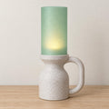 Alpine Jade Avelune lamp with a White Marble speckled base and loop handle, Light Jade cylindrical shade and soft tea light glow on a wood tabletop.
