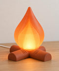 Amber Ember Emberkin lamp with an Orange translucent flame shade and Terracotta crossed-log base, illuminated on a wood tabletop with its wired cord behind.