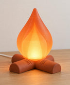 Amber Ember Emberkin lamp with an Orange translucent flame shade and Terracotta crossed-log base, illuminated on a wood tabletop with its wired cord behind.