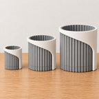 Three Linen Shadow Arvenna planters arranged from small to large on oak, showing proportional widths and the sweeping wrap over each ribbed pot.