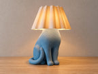 Blue seated cat lamp with white curved fluted shade, close-up three-quarter view on light oak against a pale gray wall.