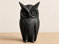 Charcoal owl sculpture, close-up front view on pale oak against an ivory wall.