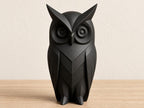 Charcoal owl sculpture, close-up front view on pale oak against an ivory wall.