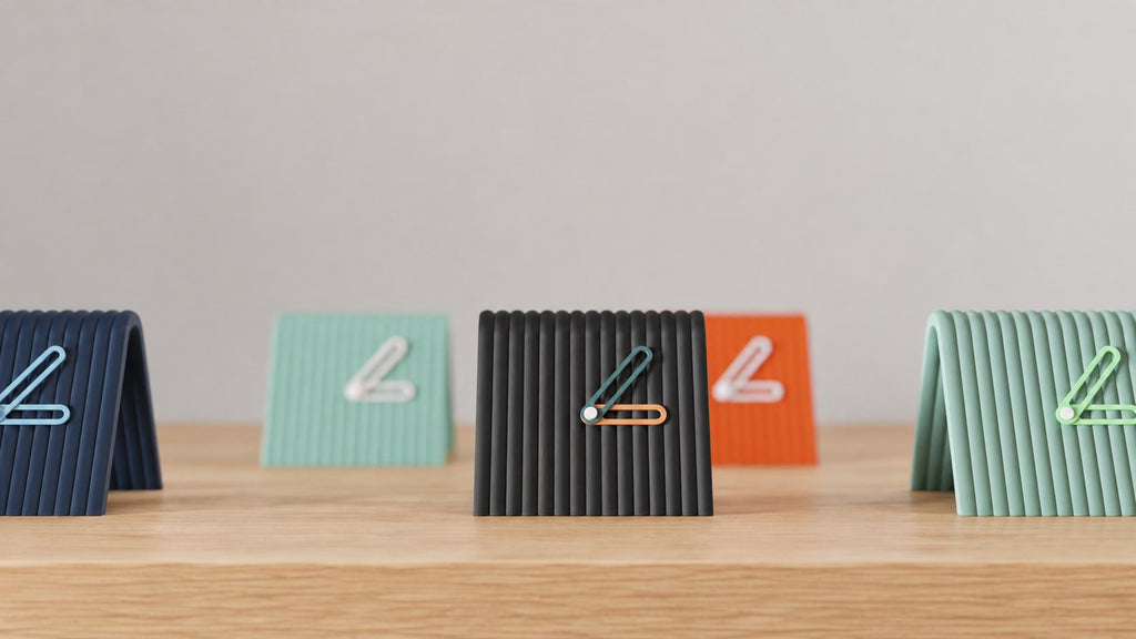 Angled close-up of Linefold Compact Desk Clock examples in contrasting colors on light oak against a gray wall.