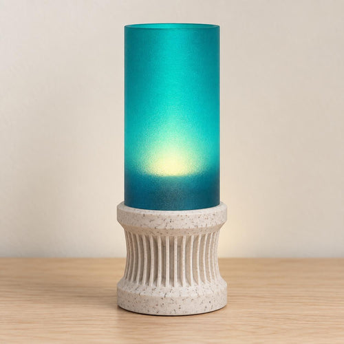 Marble Tide Alvara lamp with a White Marble speckled ribbed base and softly glowing Teal cylindrical shade on a pale wood tabletop.