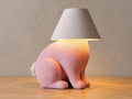 Pink rabbit lamp with white tail and smooth white shade, close-up three-quarter view on light oak against a pale gray wall.