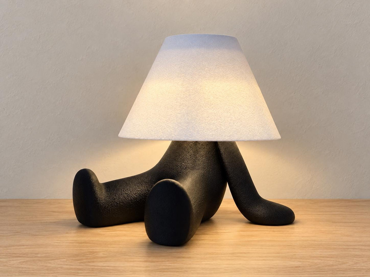Black reclining character lamp with extended legs and white smooth shade, close-up three-quarter view on light oak against a pale gray wall.