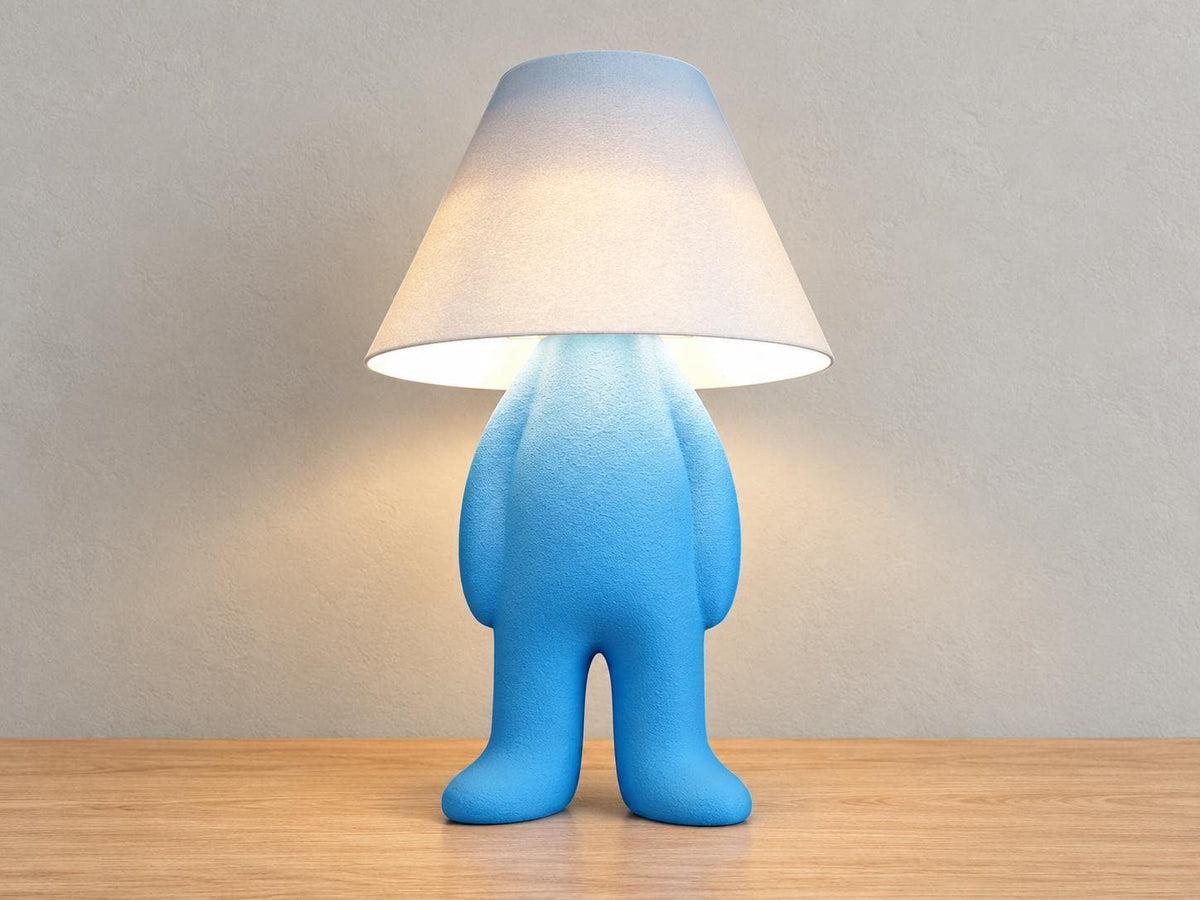 Blue upright character lamp with arms down and white smooth shade, close-up three-quarter view on light oak against a pale gray wall.