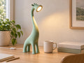 Pale green 17 inch Canopy giraffe lamp with wired USB light beside a mug, books, and leafy plant in a warm home office