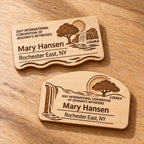 Two Lusaka mini natural-birch engraved badge designs, slot-free and laid separately on a light oak tabletop; each shows sample personalization.