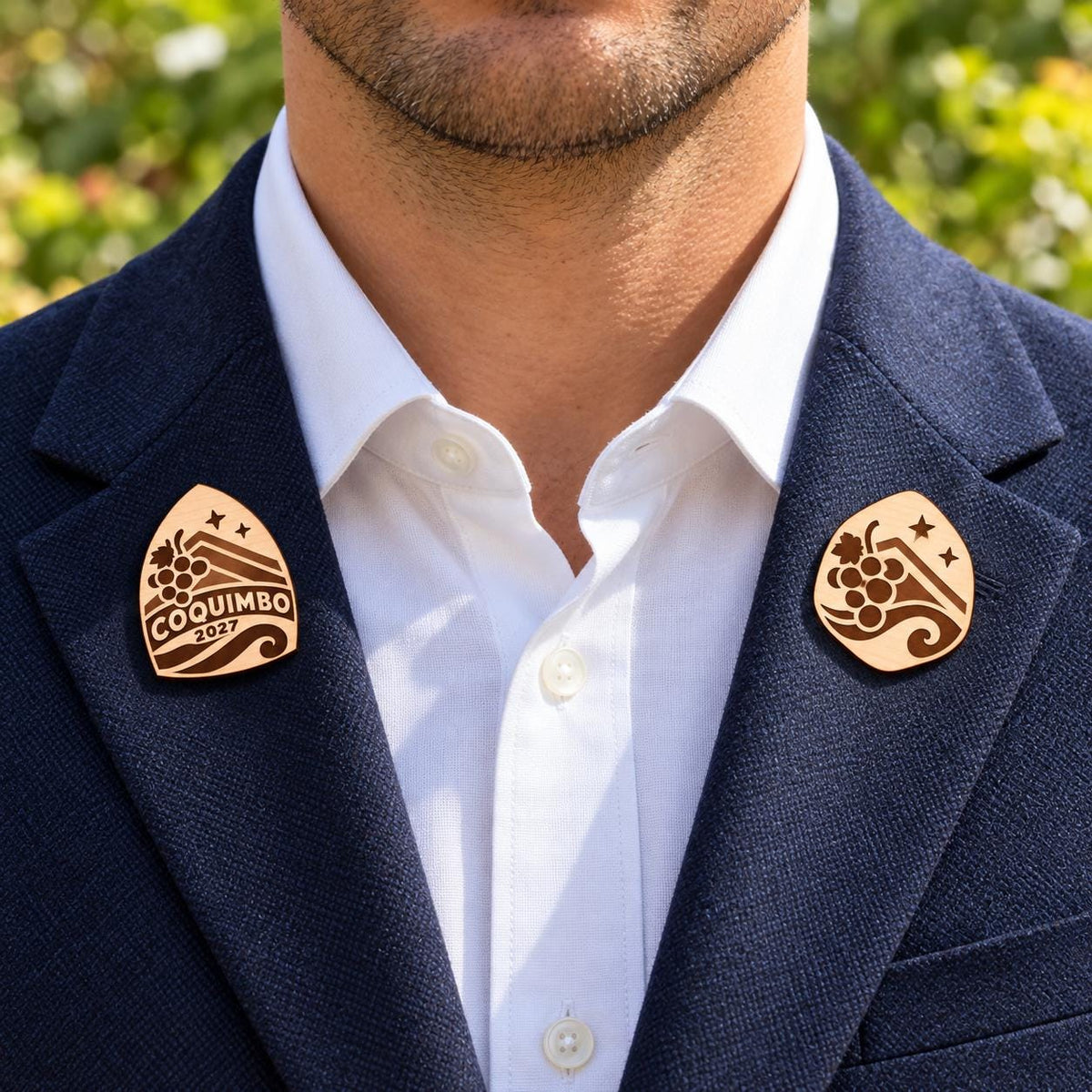 Two Coquimbo engraved birch convention pin designs on the lapels of a navy man's suit outdoors.