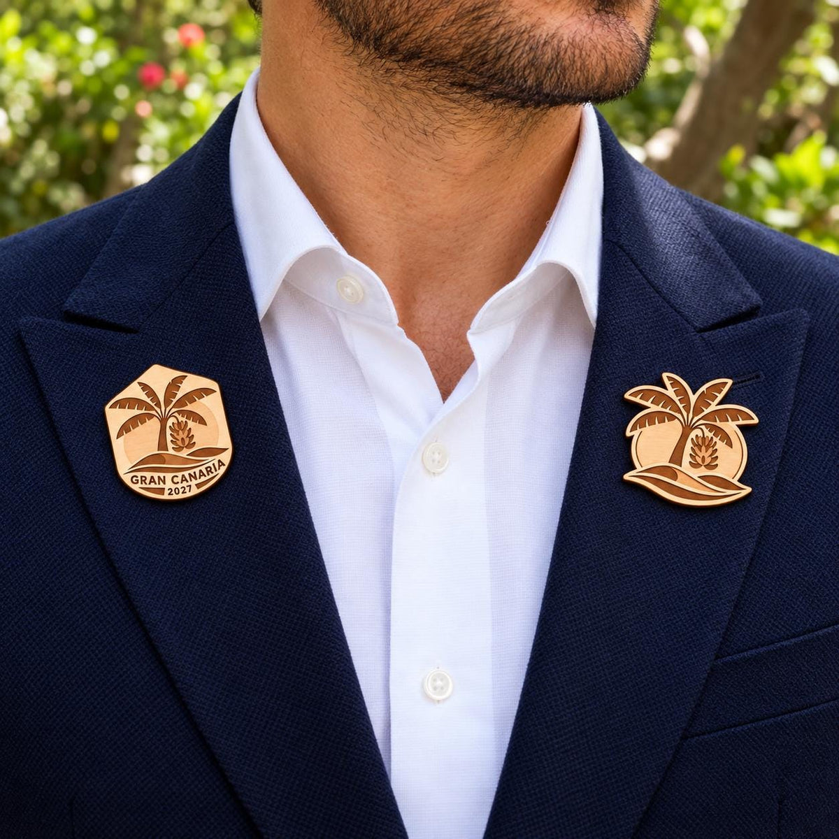 Two Gran Canaria engraved birch convention pin designs on the lapels of a navy man's suit outdoors.