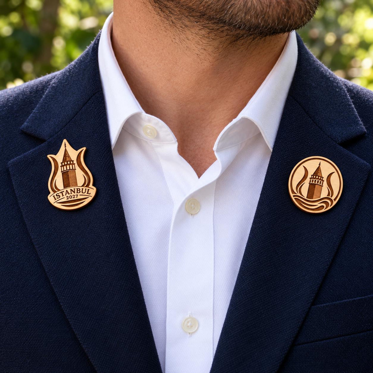Two Istanbul engraved birch convention pin designs on the lapels of a navy man's suit outdoors.