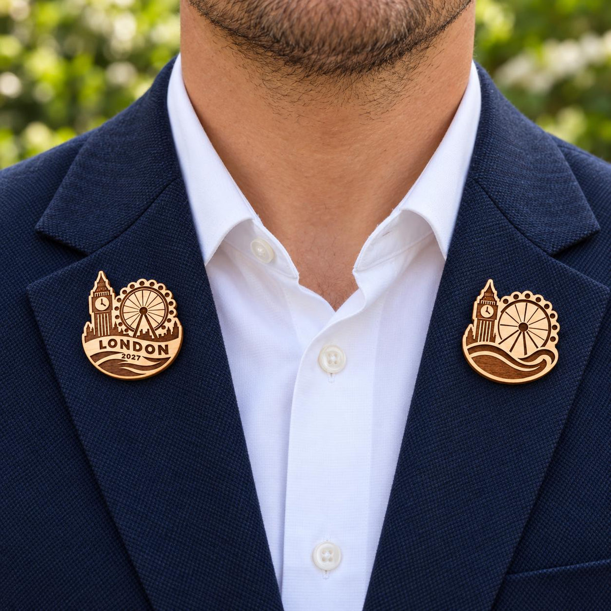 Two London engraved birch convention pin designs on the lapels of a navy man's suit outdoors.