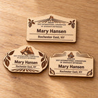 Three Abidjan mini natural-birch engraved badge designs, slot-free and laid separately on a light oak tabletop; each shows sample personalization.