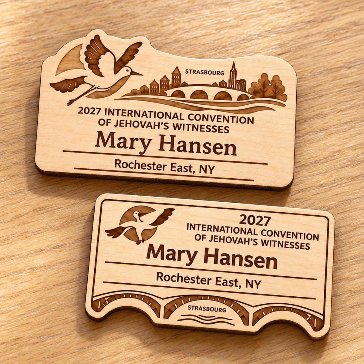 Two Strasbourg mini natural-birch engraved badge designs, slot-free and laid separately on a light oak tabletop; each shows sample personalization.