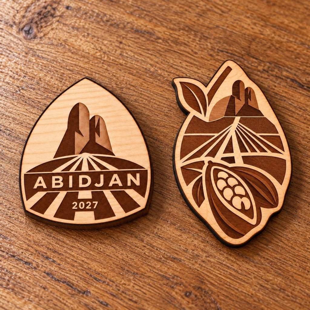 Two Abidjan engraved birch convention pin designs lying face-up on a wooden table in natural light.