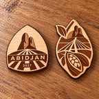 Two Abidjan engraved birch convention pin designs lying face-up on a wooden table in natural light.
