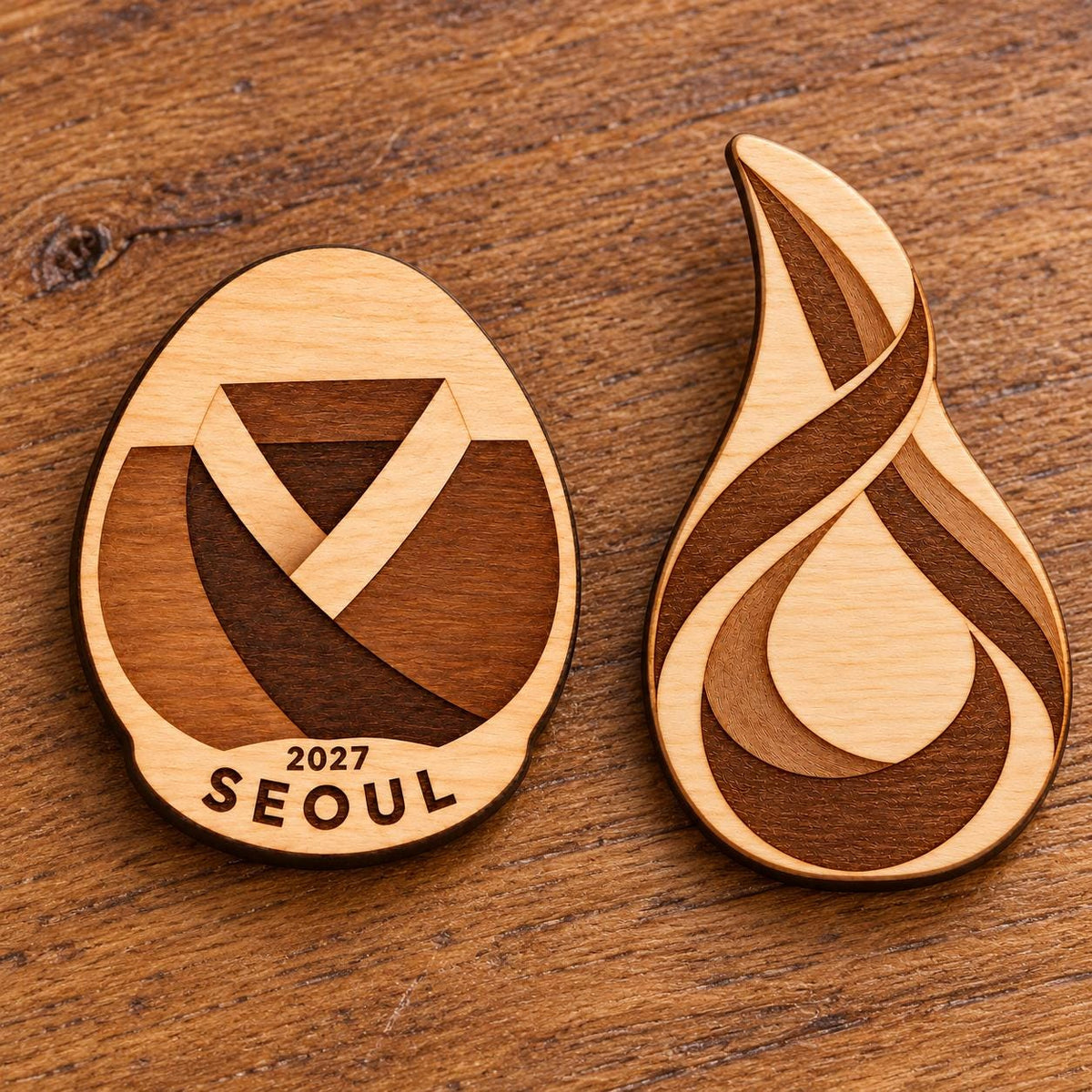 Two Seoul engraved birch convention pin designs lying face-up on a wooden table in natural light.