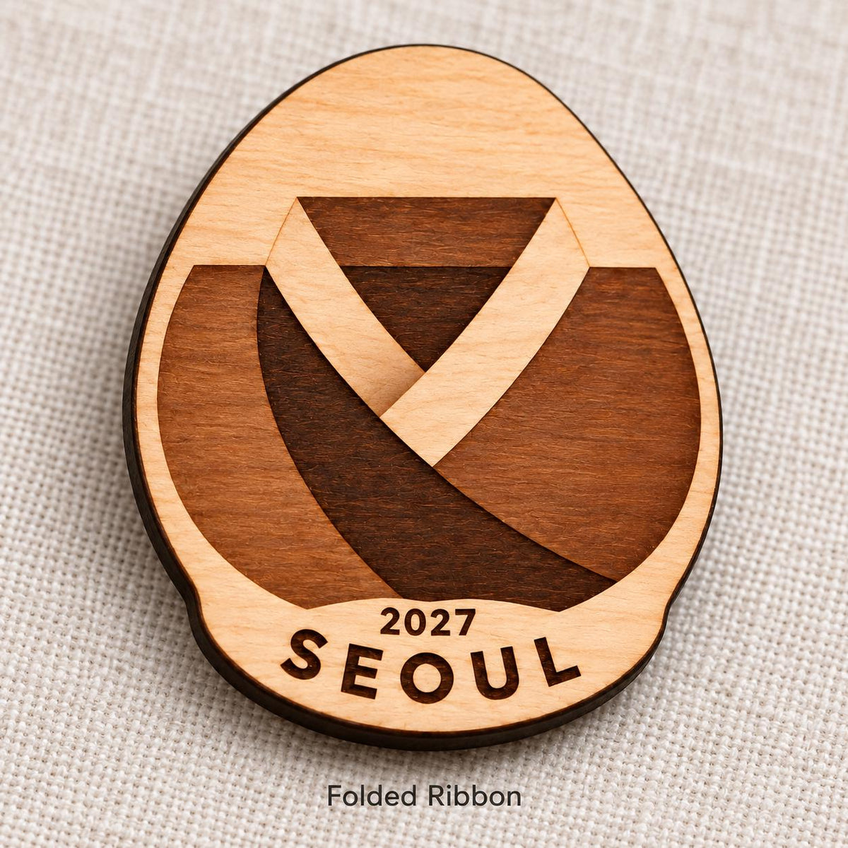 Close-up of Seoul Folded Ribbon engraved birch pin on linen, with design name caption.