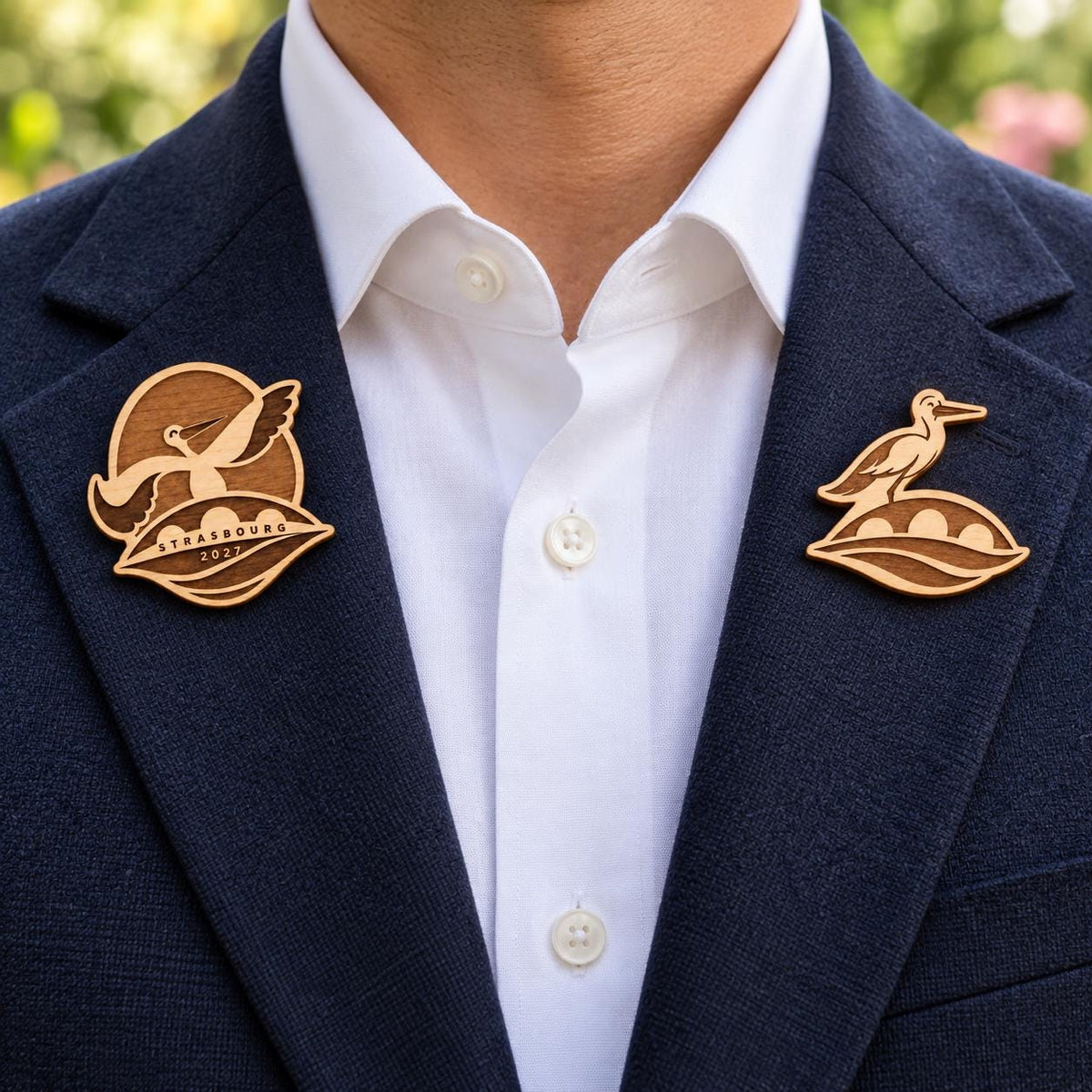 Two Strasbourg engraved birch convention pin designs on the lapels of a navy man's suit outdoors.