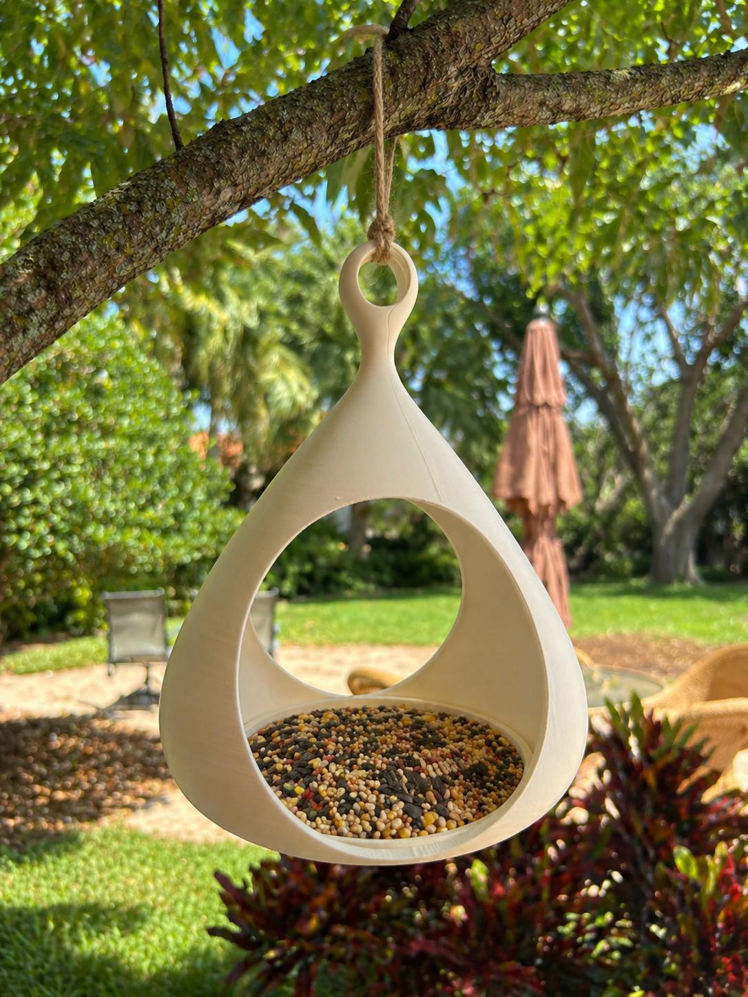 Cream teardrop-shaped bird feeder hanging from a leafy backyard tree.
